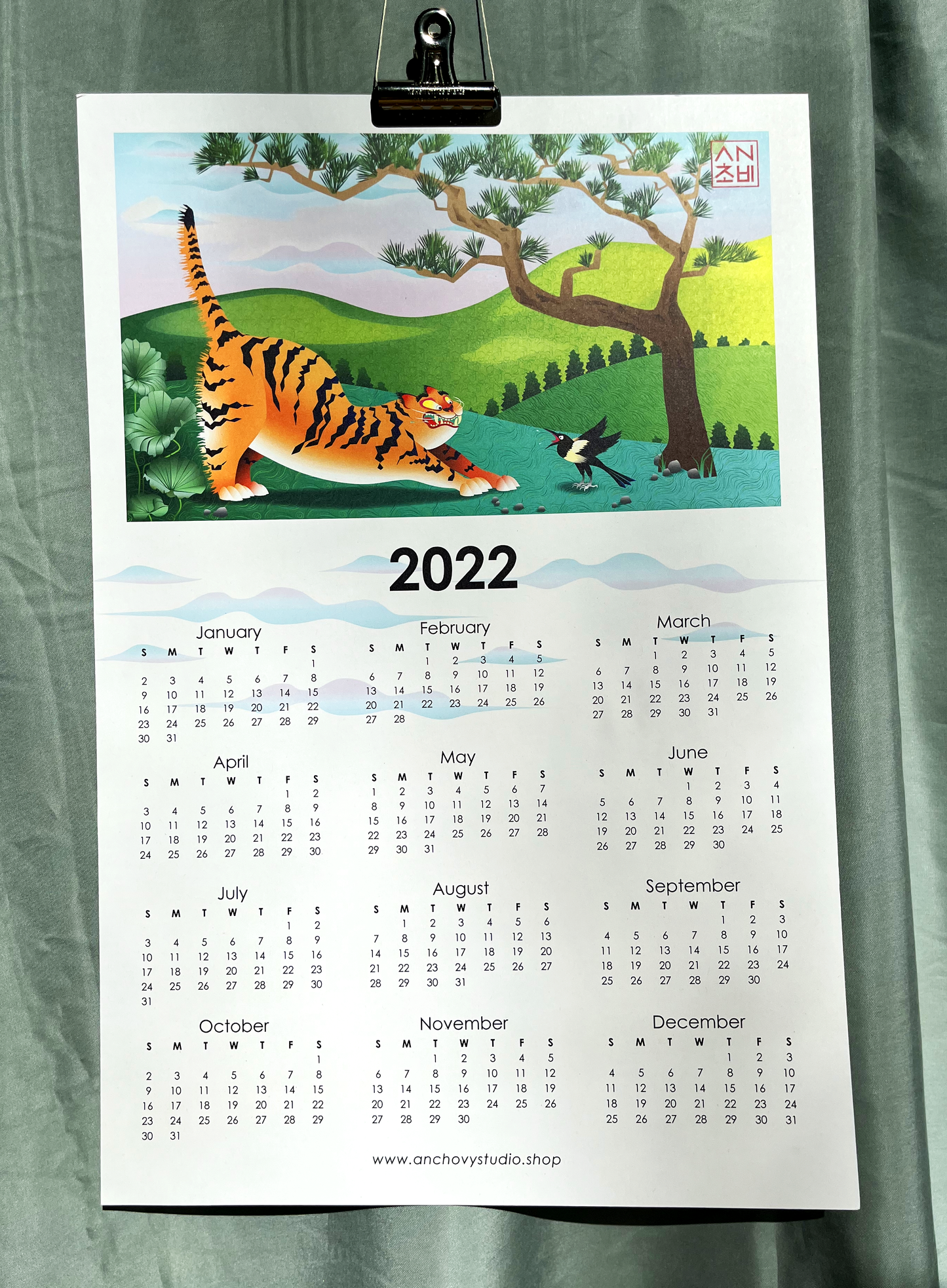 12x18 Color 2022 Calendar Year of the Tiger Print for home, apartment, and room decor. Illustration printed in full color with all 12 months of the year. Inspired by Korean minhwa painting, a magpie scares the wits out of a tiger in a lush landscape of lotus, pine, and rolling hills.